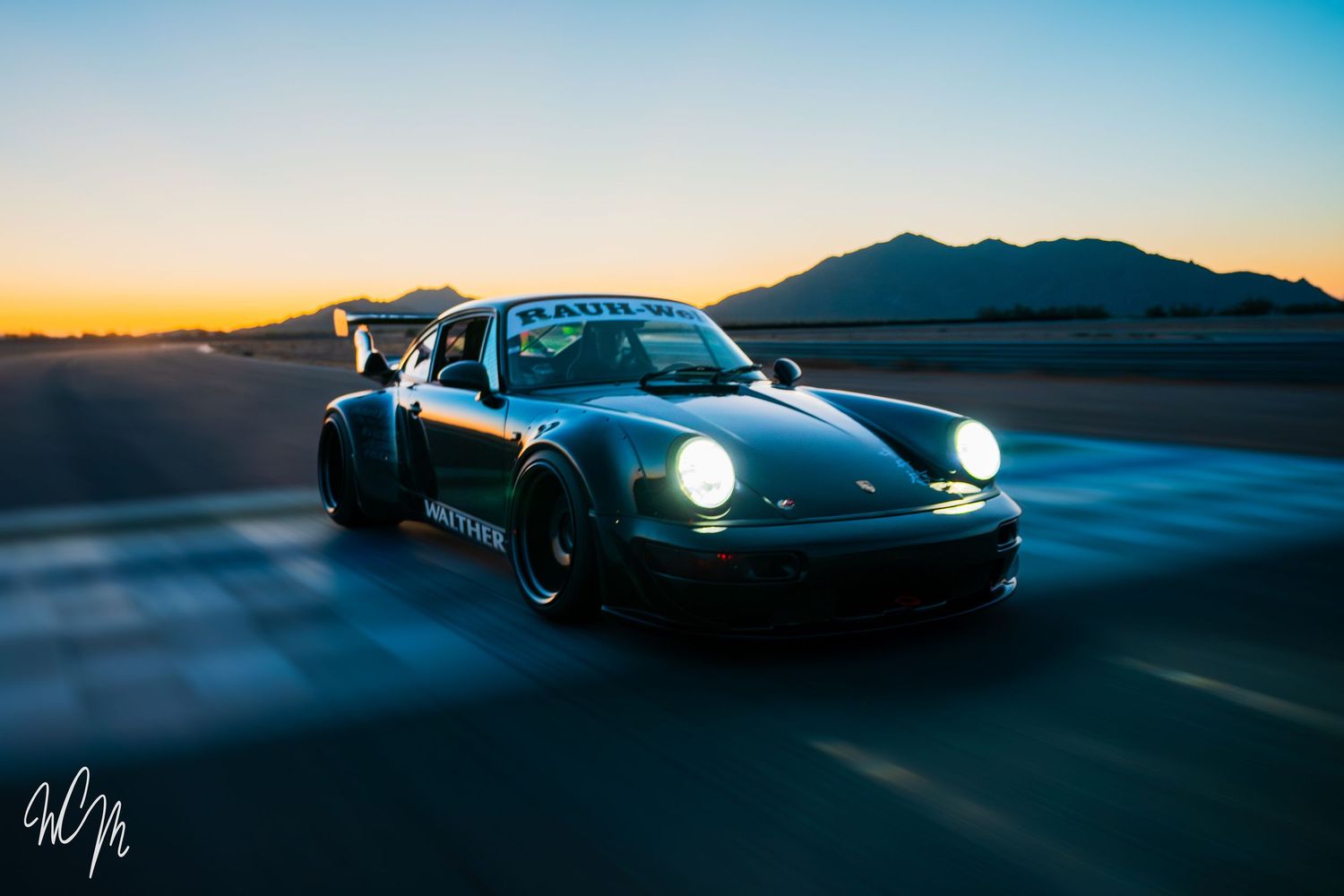 Custom Porsche 911 with widebody kit driving at sunset with mountain silhouettes.