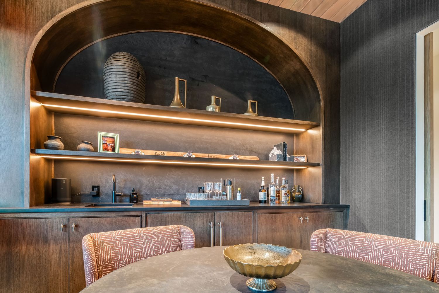Dark wood built-in shelving with integrated lighting displays decorative items in a modern home bar setup.