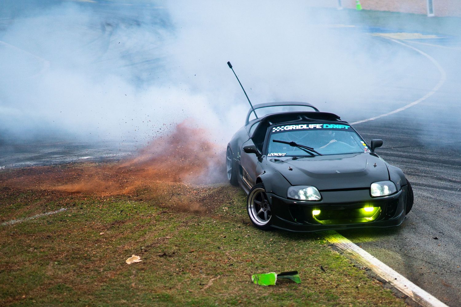 Black drift car performing smoky burnout on track with green underglow lights and tire smoke filling the air.