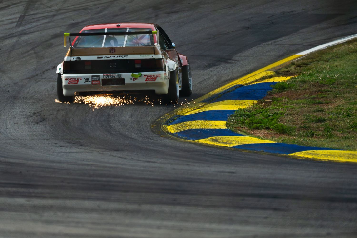 Race car sparking on track corner during motorsport event, captured at low angle with dramatic lighting.