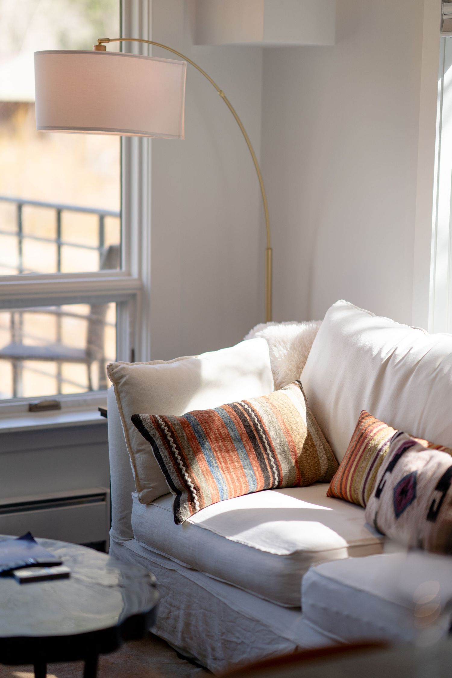 Sunlit white sofa with colorful striped pillows beside an arched gold floor lamp near a bright window.