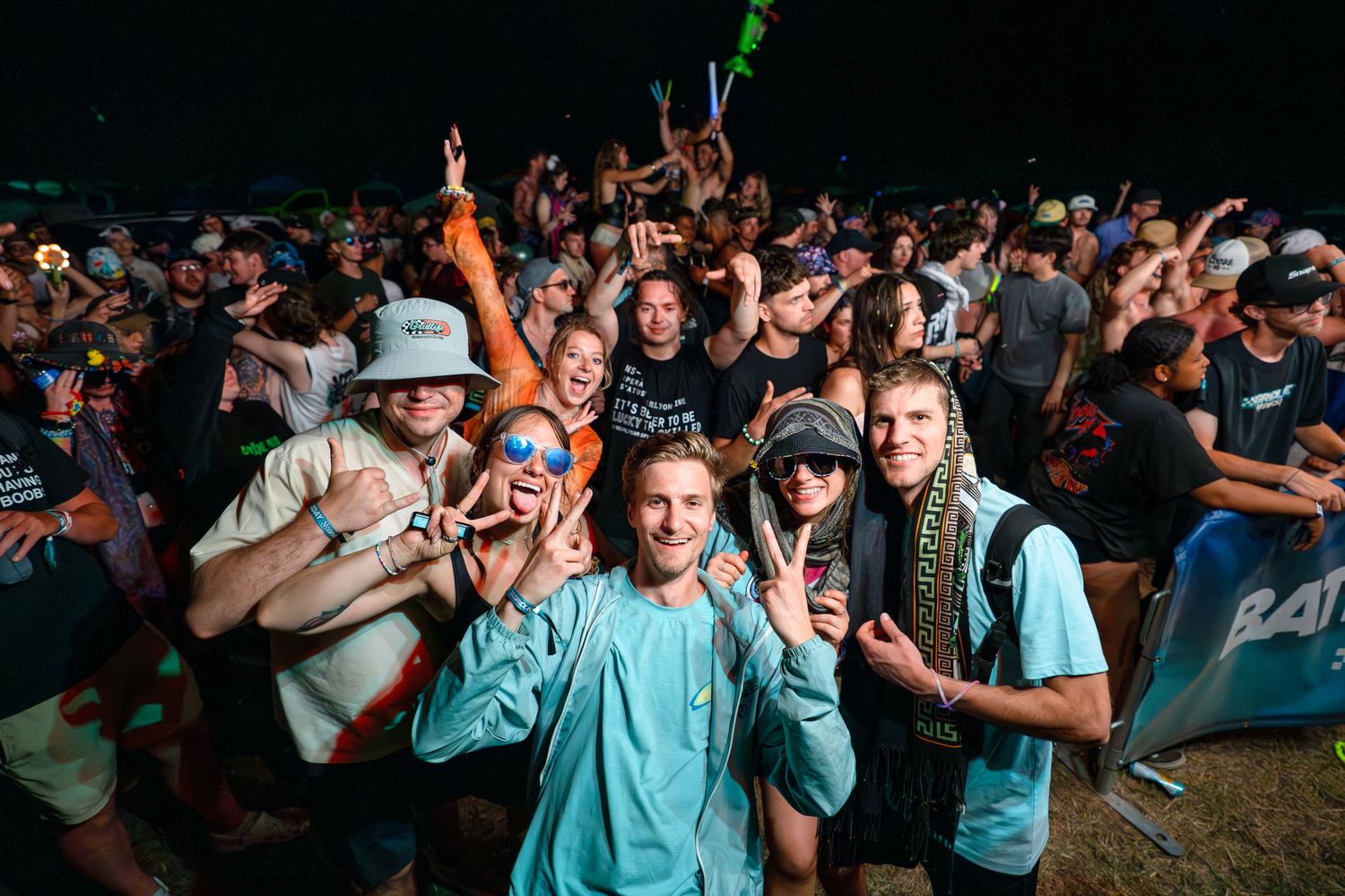 Crowd of young festival-goers celebrating at night, smiling and posing for camera at outdoor music event.