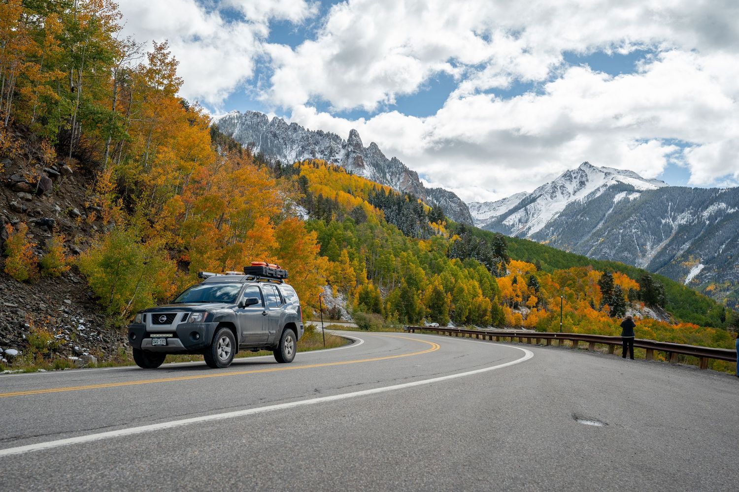 A scenic mountain road lined with golden autumn trees and snow-capped peaks under a cloudy sky.