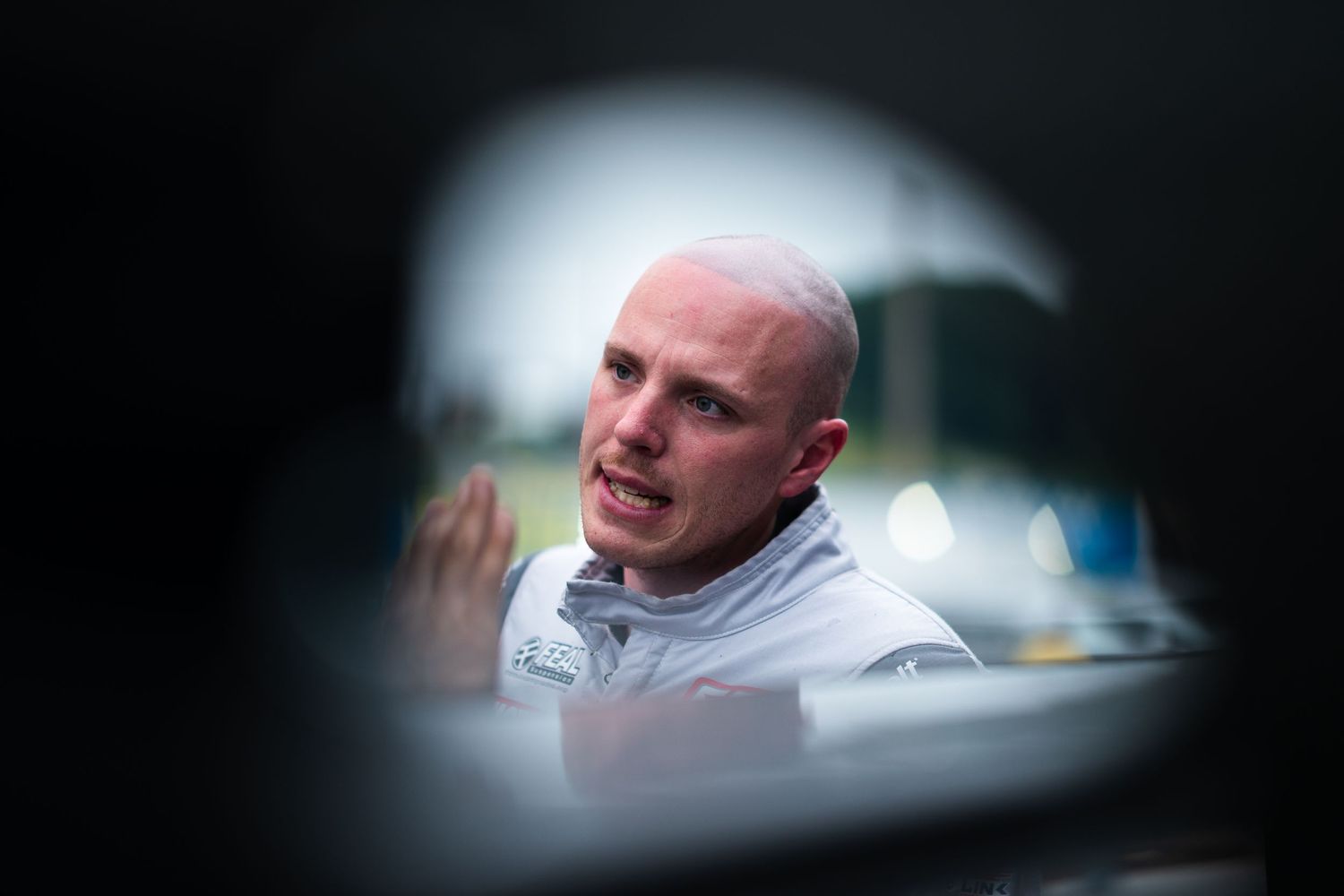 A bald racing driver in a white suit speaks candidly, photographed through a car window reflection at a motorsport event.
