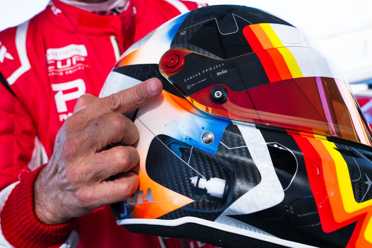 Close-up of a colorful racing helmet with red, black and yellow design being held by hands in a red racing suit.