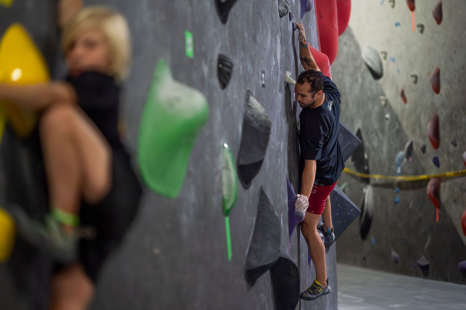 Two climbers tackle a colorful indoor bouldering wall with green and yellow holds in a climbing gym, one man gripping the wall mid-climb.
