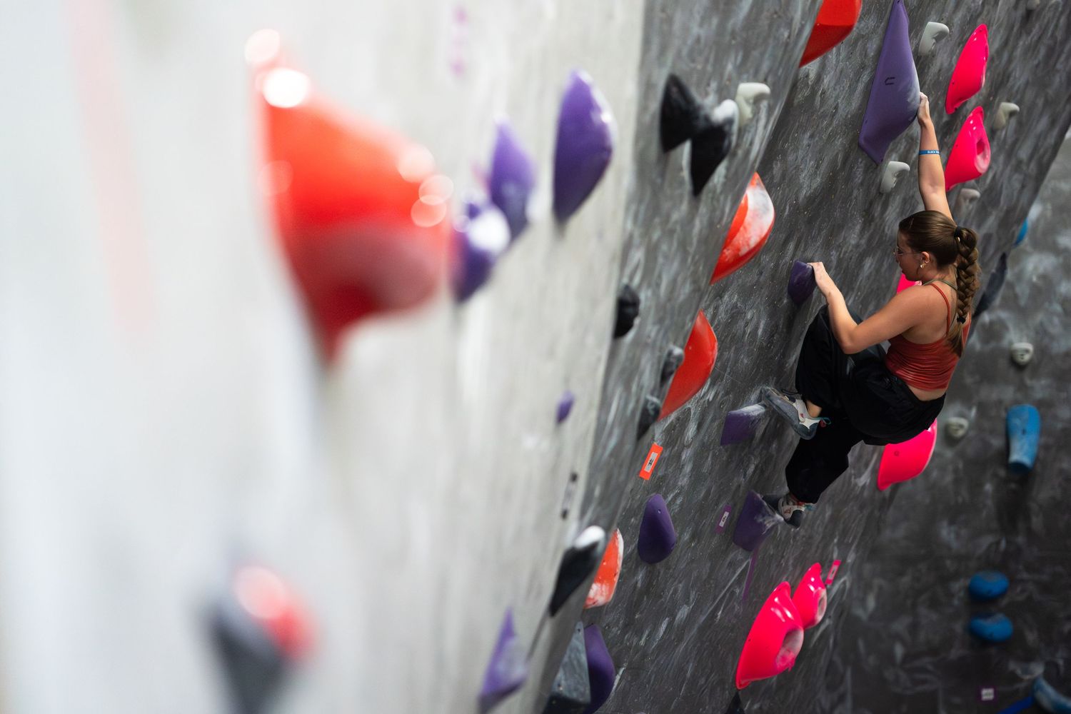 Woman with a ponytail climbs an indoor rock wall covered in purple, red and orange holds.