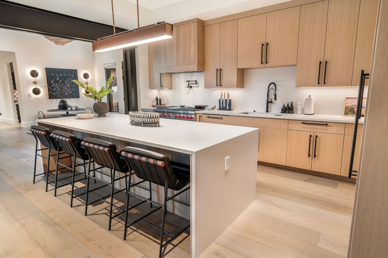 Modern kitchen interior with light wood cabinets, quartz countertops, and black bar stools along an island peninsula.