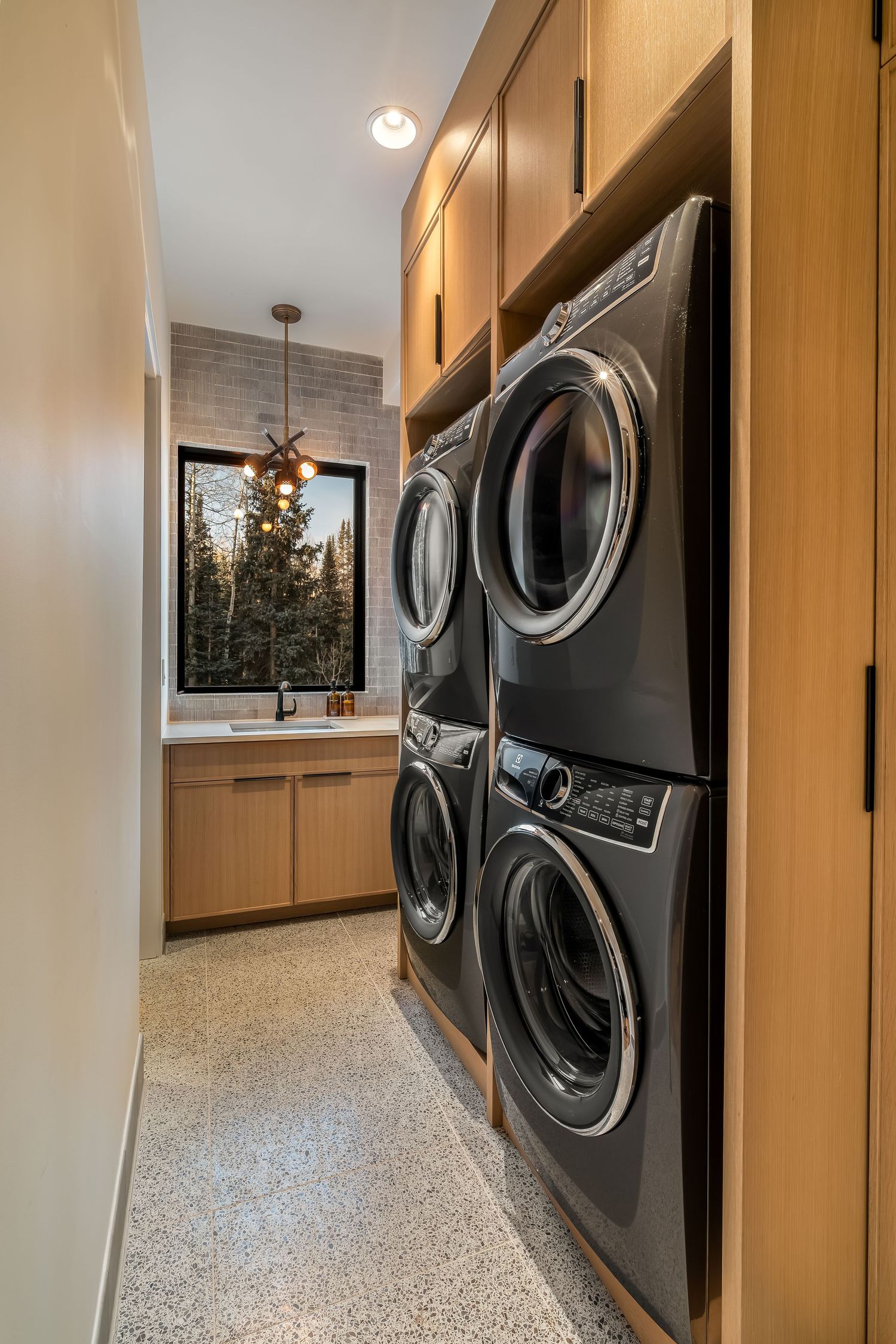 Modern laundry room with stacked washers and dryers and window overlooking pine trees.