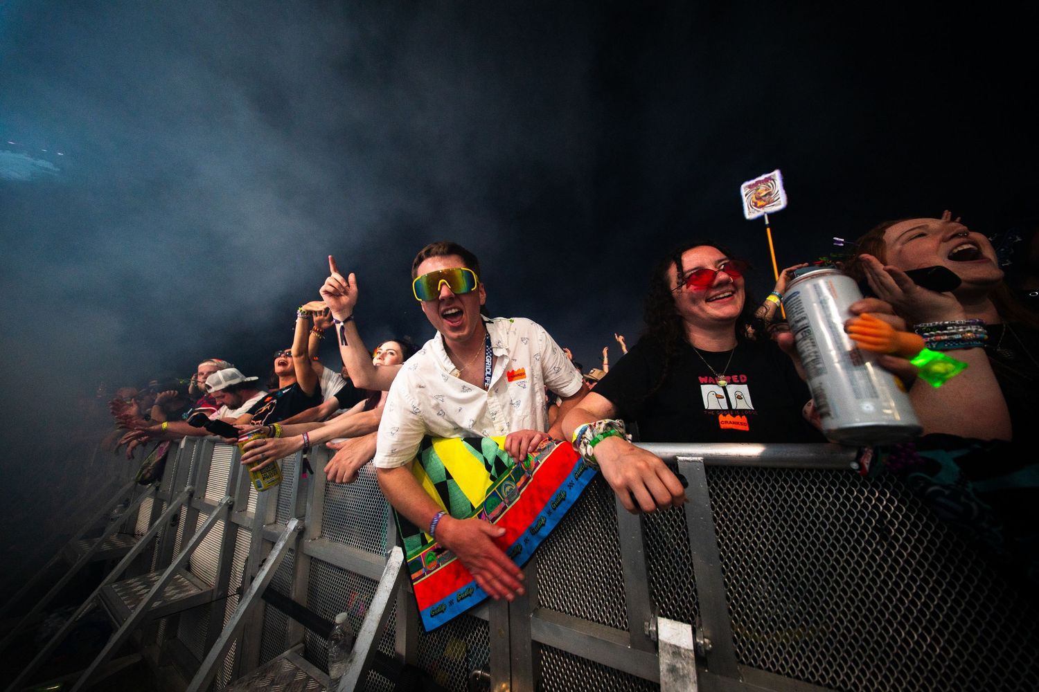 Excited concert crowd cheering behind barrier at outdoor music festival with colorful outfits and smoky atmosphere.