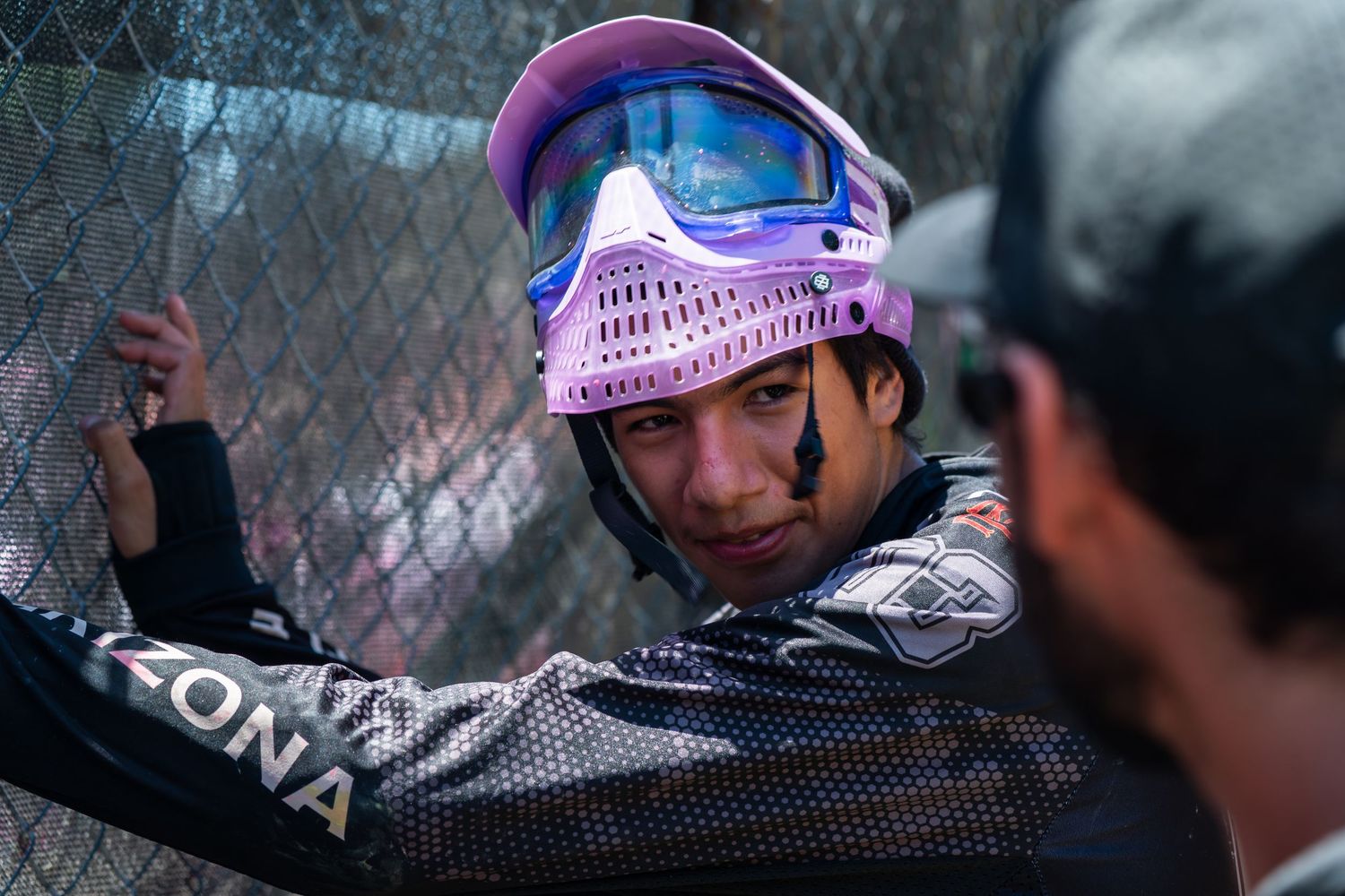 Paintball player wearing pink protective mask leaning against fence at outdoor tournament.