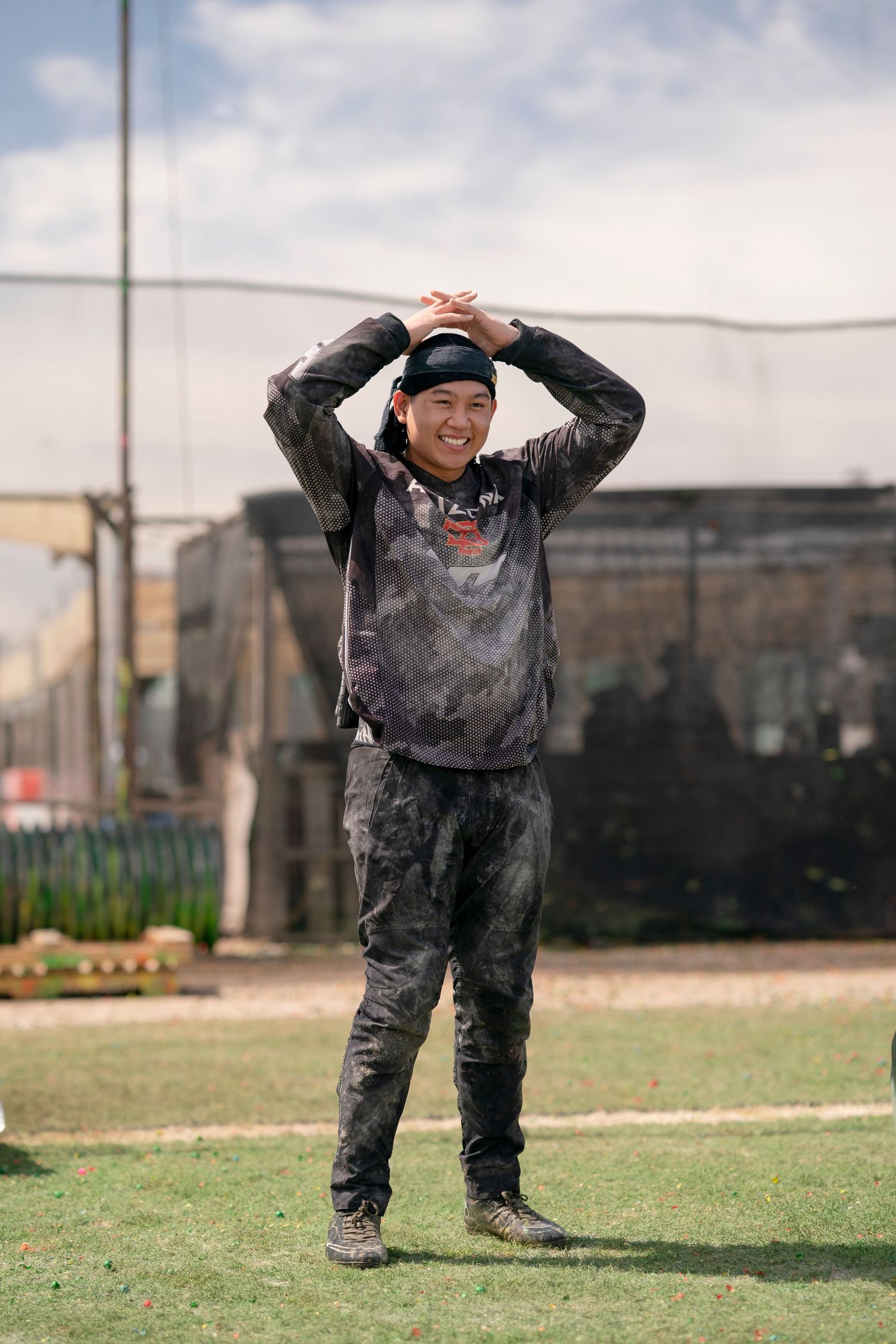 Player in paintball gear celebrates on outdoor field with buildings in background.