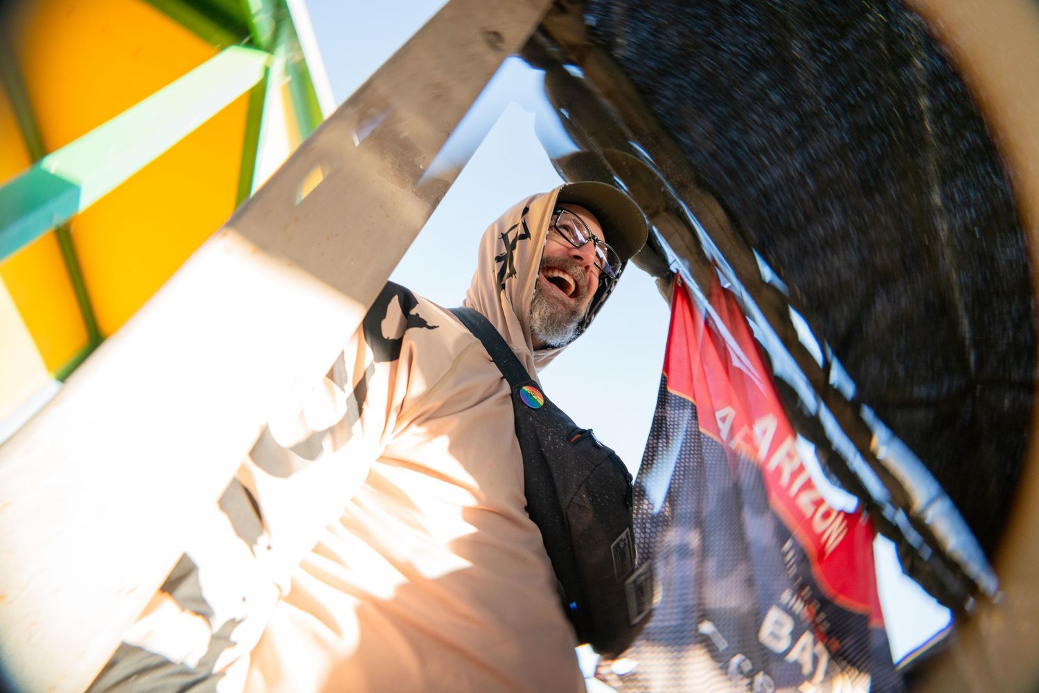 A smiling man in a hoodie and sunglasses photographed from below against a bright blue sky at an outdoor event.