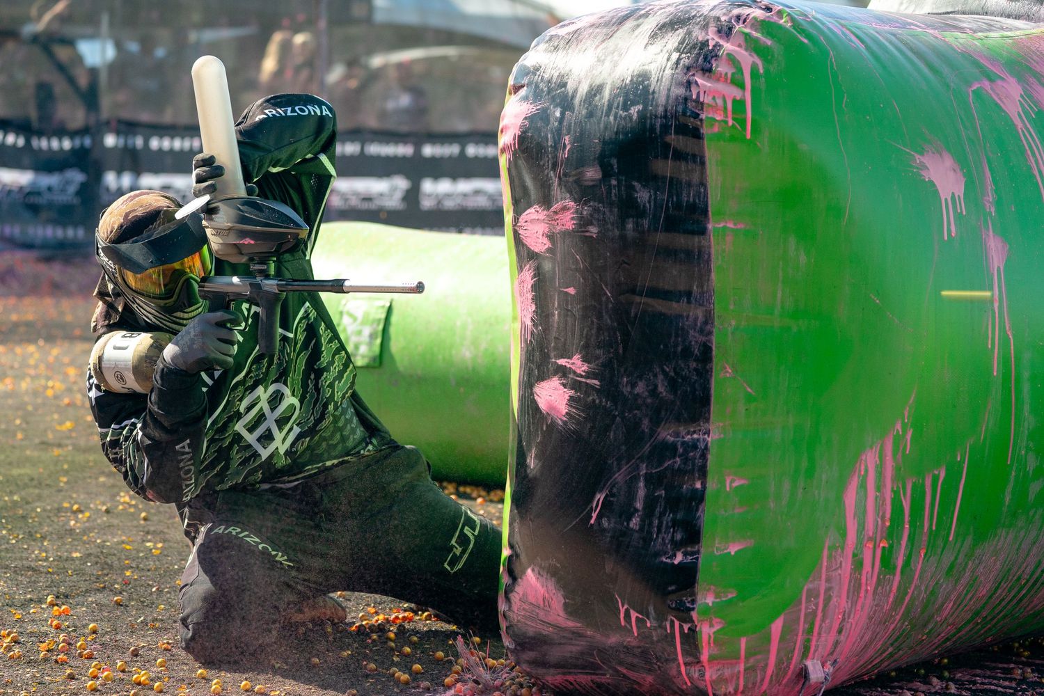 Two paintball players crouch behind a large green inflatable bunker during an intense outdoor paintball match.