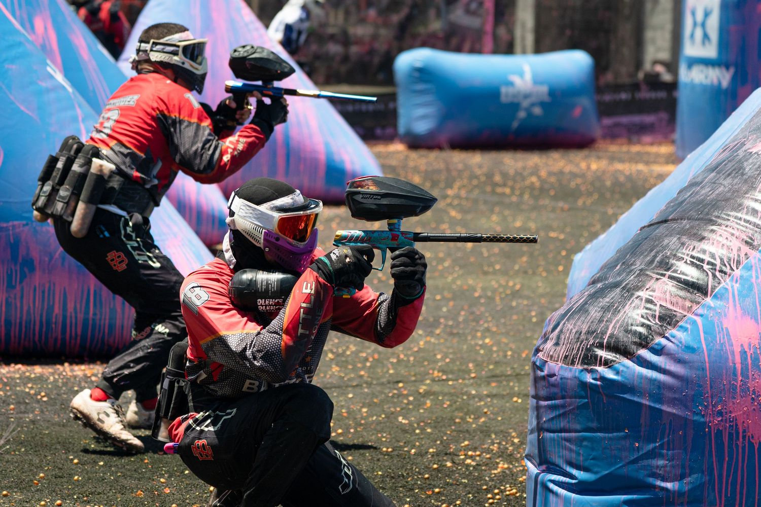 Two red-uniformed paintball players crouch behind inflatable bunkers during competitive tournament game.
