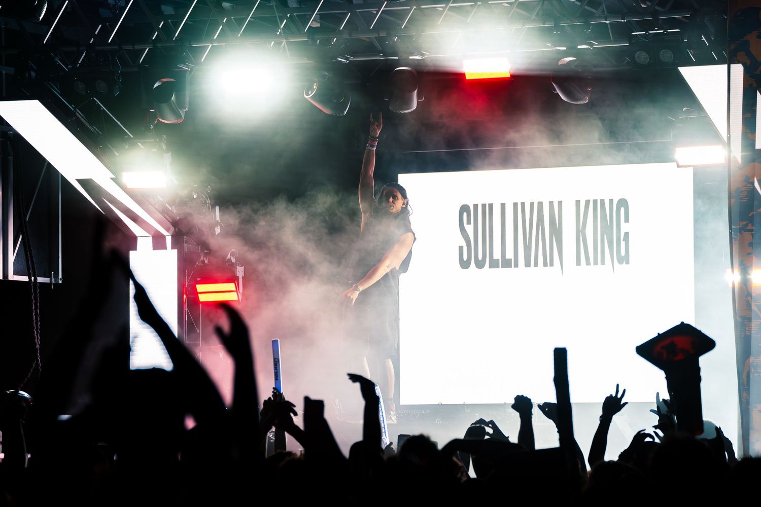 Sullivan King performs on a smoky festival stage with dramatic lighting as an energetic crowd raises their hands.