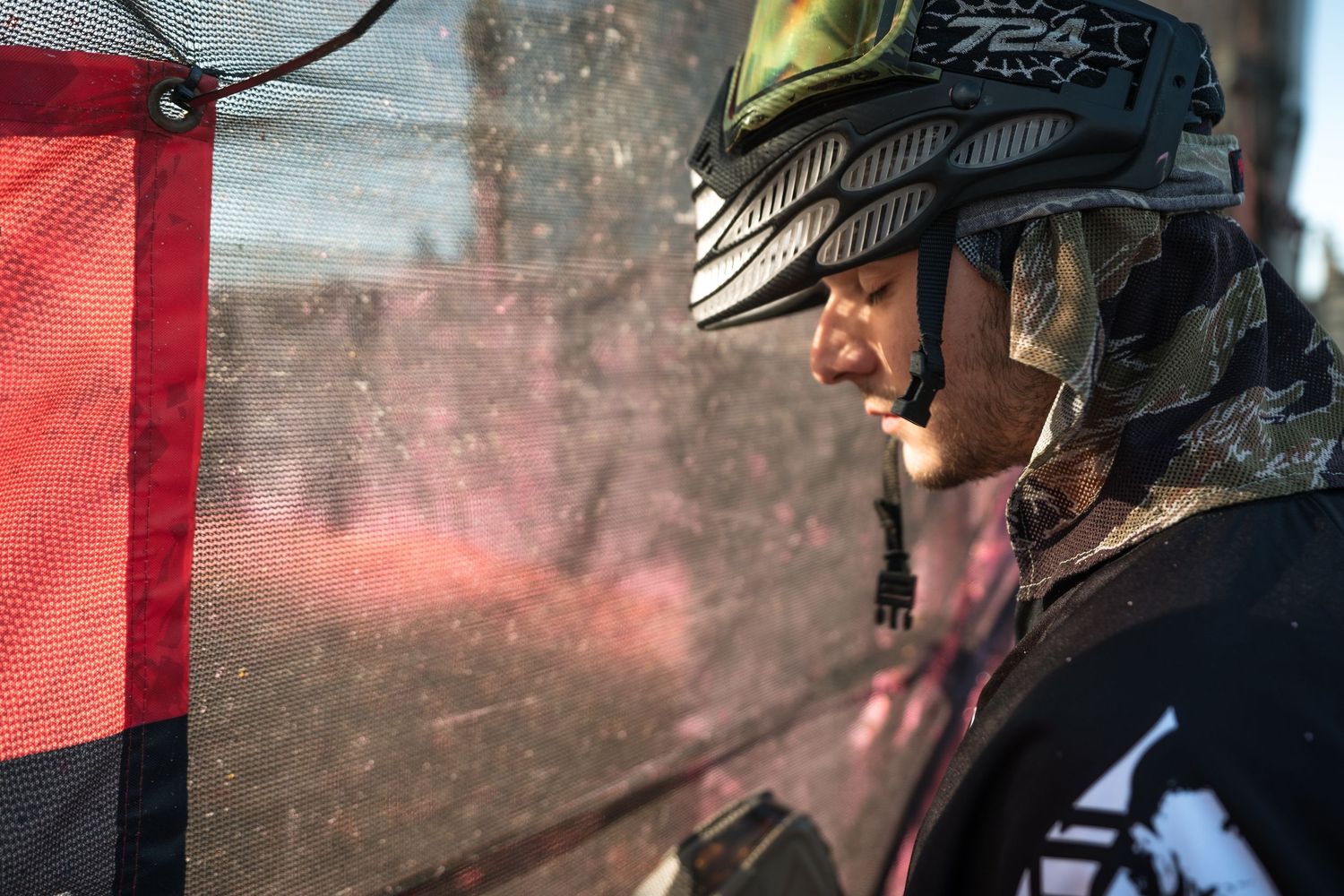 A paintball player in protective helmet and gear peers around a mesh barrier during an intense match.