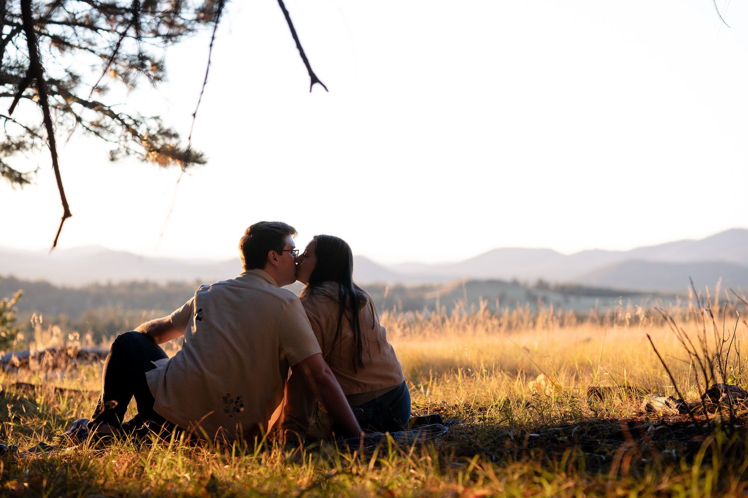 Couple sharing a kiss while sitting in a golden meadow with distant mountains and a soft sunset glow.