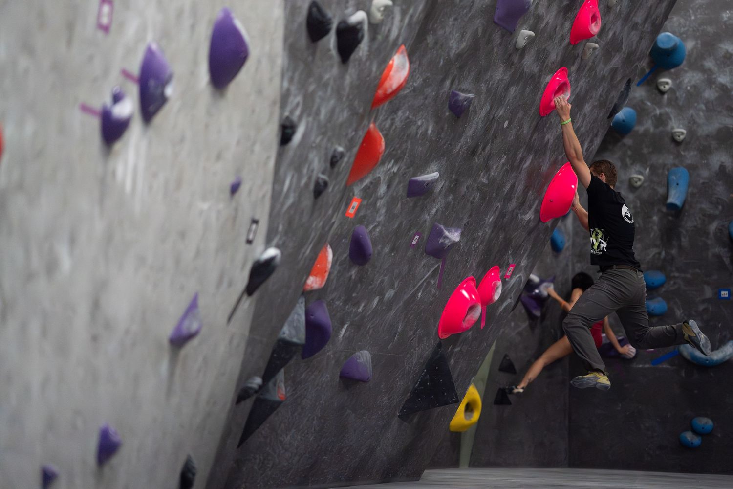 Climber in a black shirt stretches toward red and orange holds on a steep indoor bouldering wall.