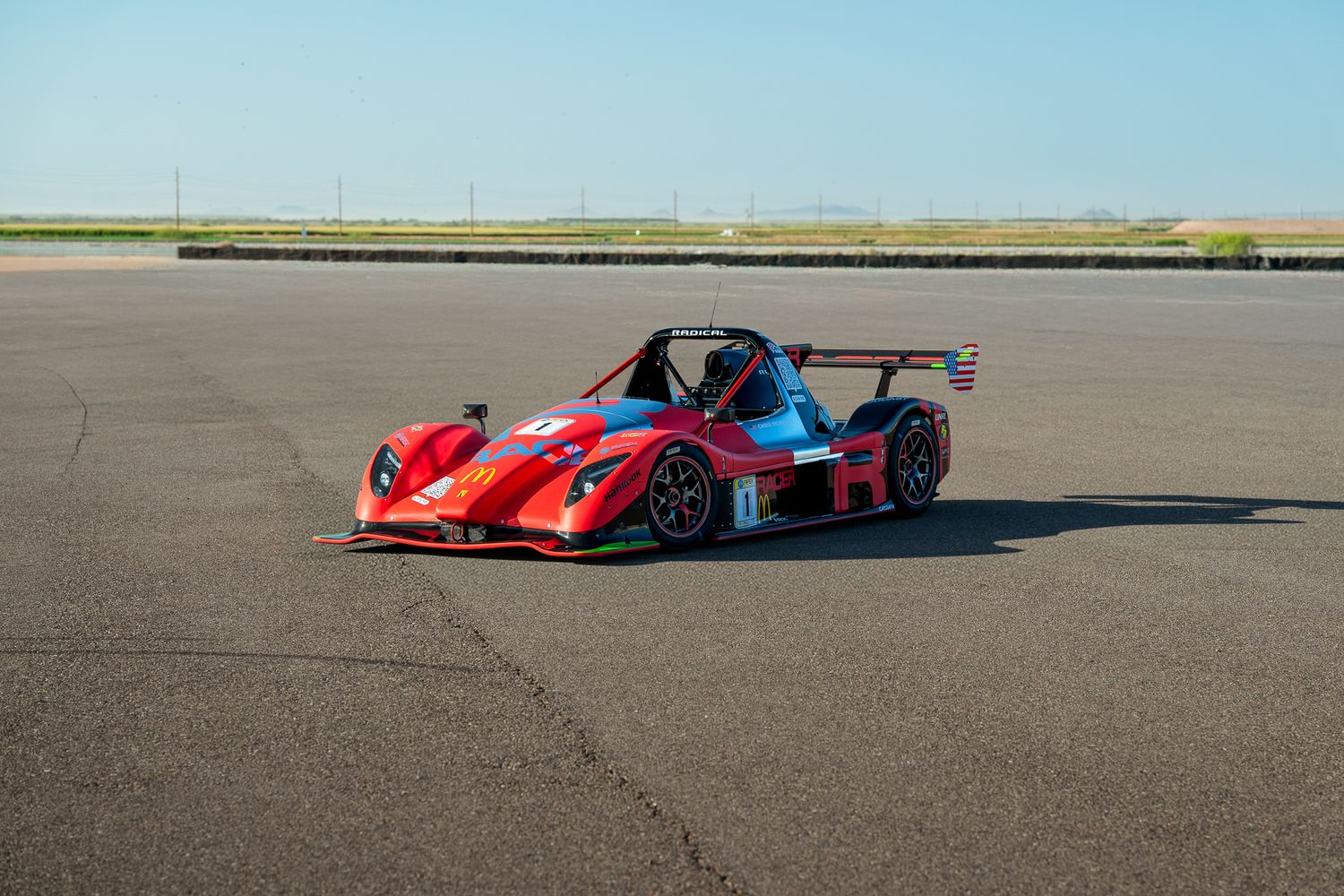 Red prototype race car with aerodynamic bodywork parked on empty tarmac under clear blue sky.