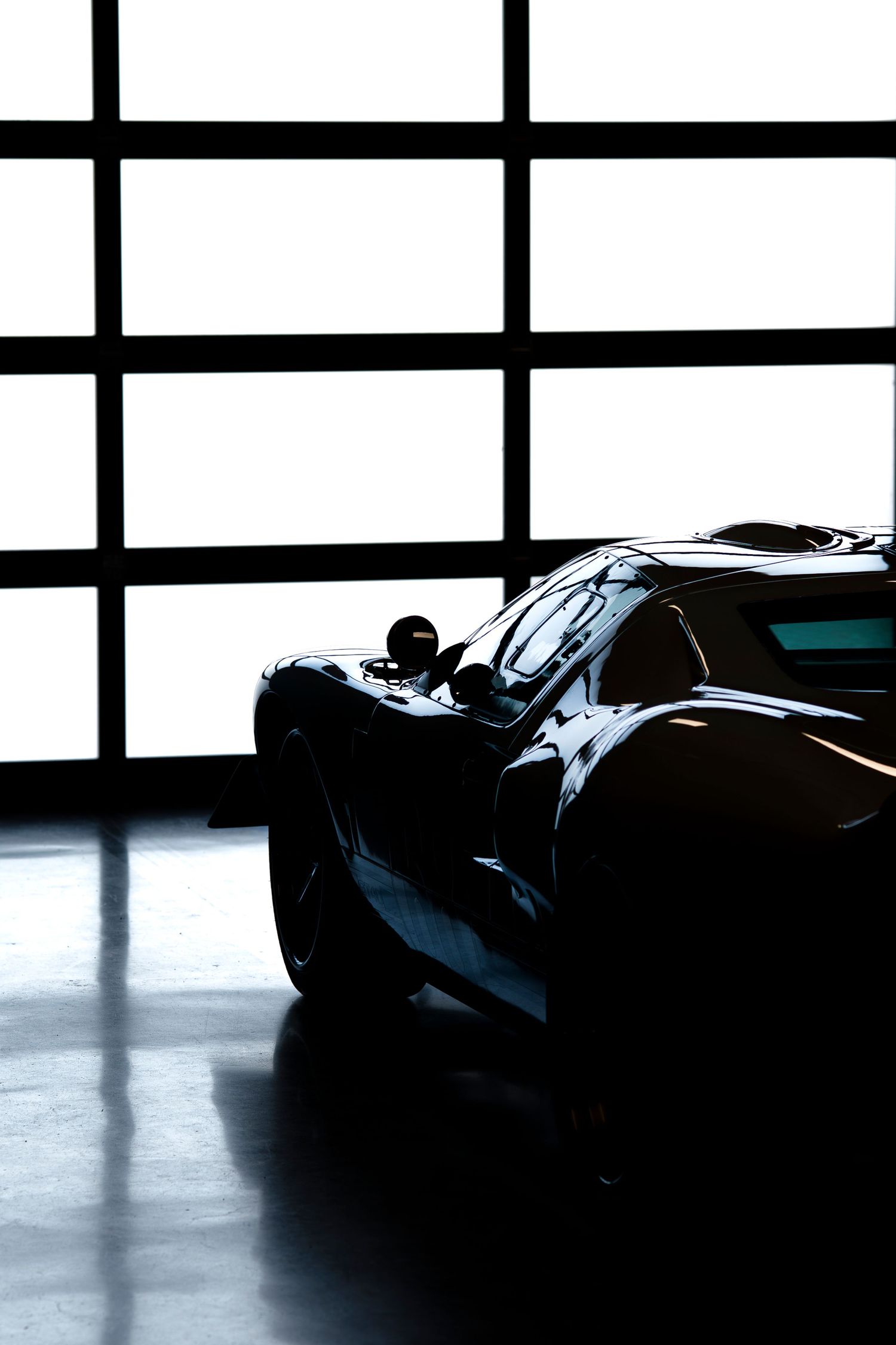 Sleek black sports car silhouette in dramatic lighting against geometric window patterns in modern showroom.