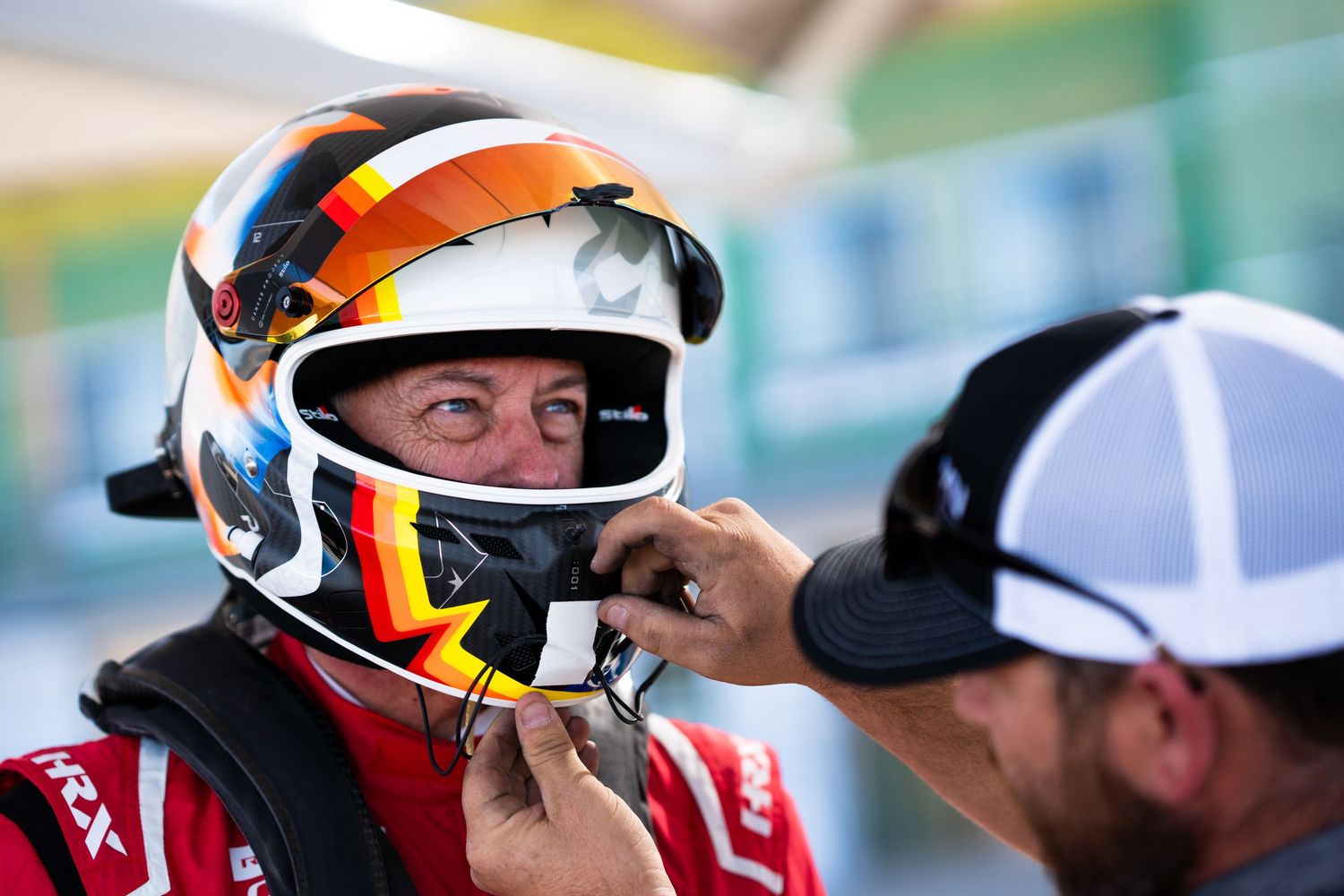 Racing driver wearing a helmet with orange and white accents looks to the side during a motorsport event.