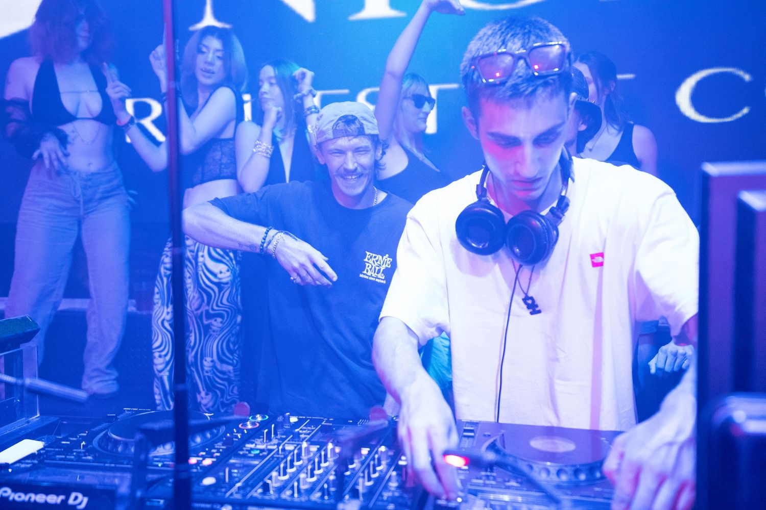 DJs perform at a nightclub with blue lighting and electronic music equipment on stage.