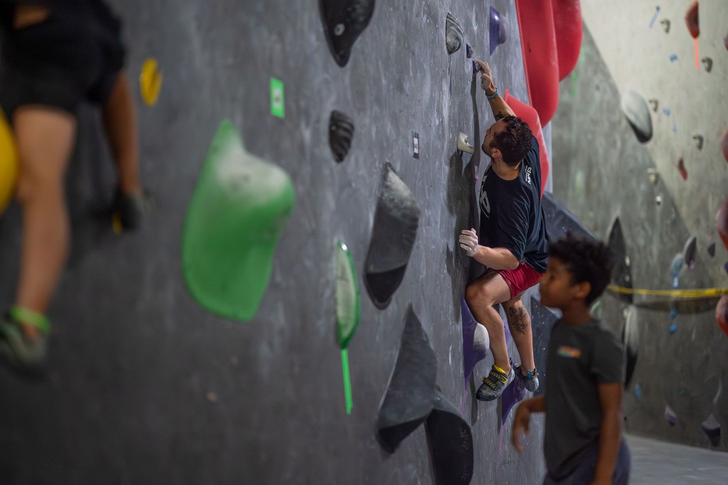 Child climber ascends a textured indoor bouldering wall with green and gray holds as a boy in a gray shirt looks on.