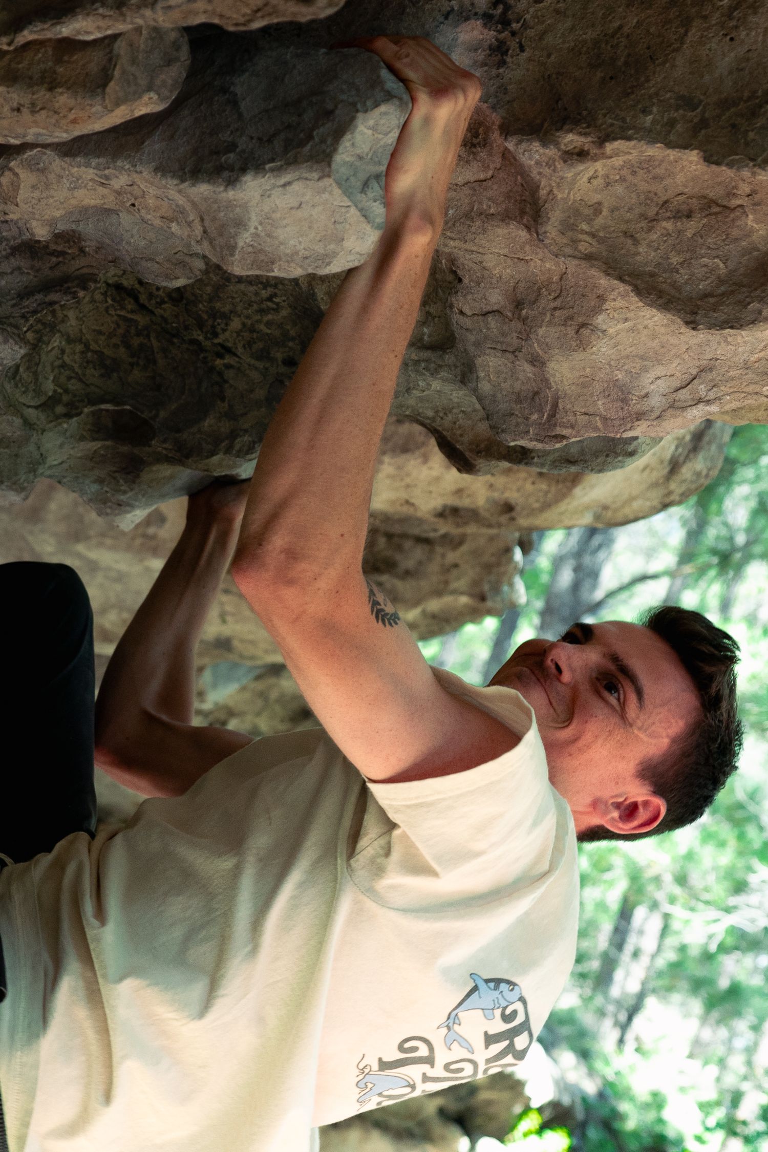 A man in a light t-shirt reaches overhead while bouldering on a rugged, overhanging rock face with green trees in the background.