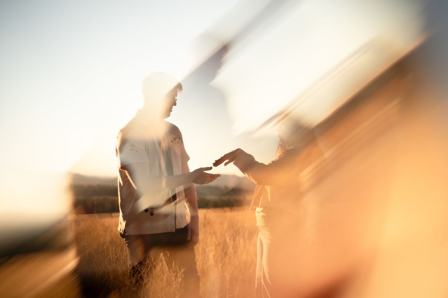 Blurred double-exposure photo of two people reaching to hold hands in a golden wheat field at sunset with warm, dreamy light.