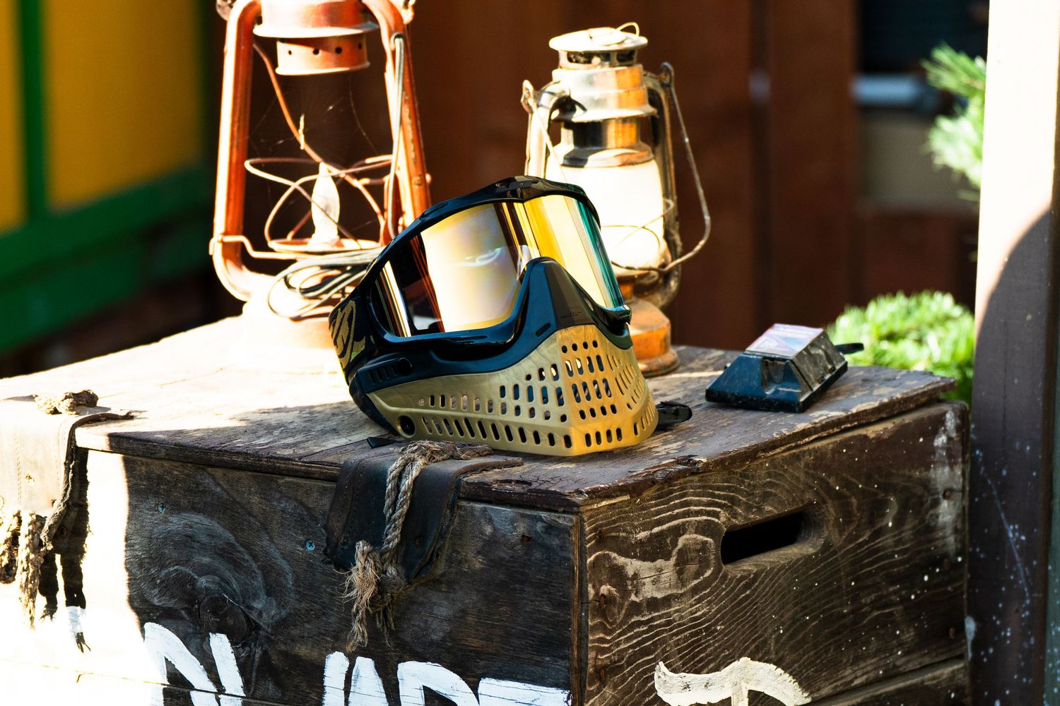 Paintball mask and vintage lanterns displayed on a wooden crate outdoors at a paintball arena.
