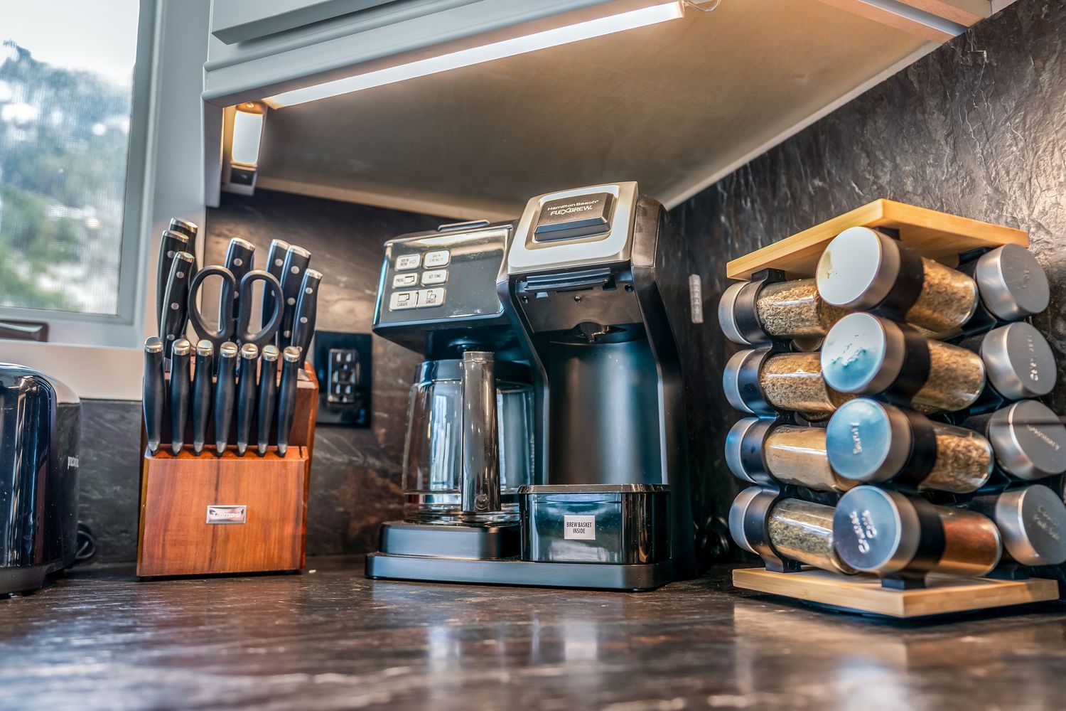 Kitchen counter with knife block, coffee maker, and spice rack organizer on dark countertop.