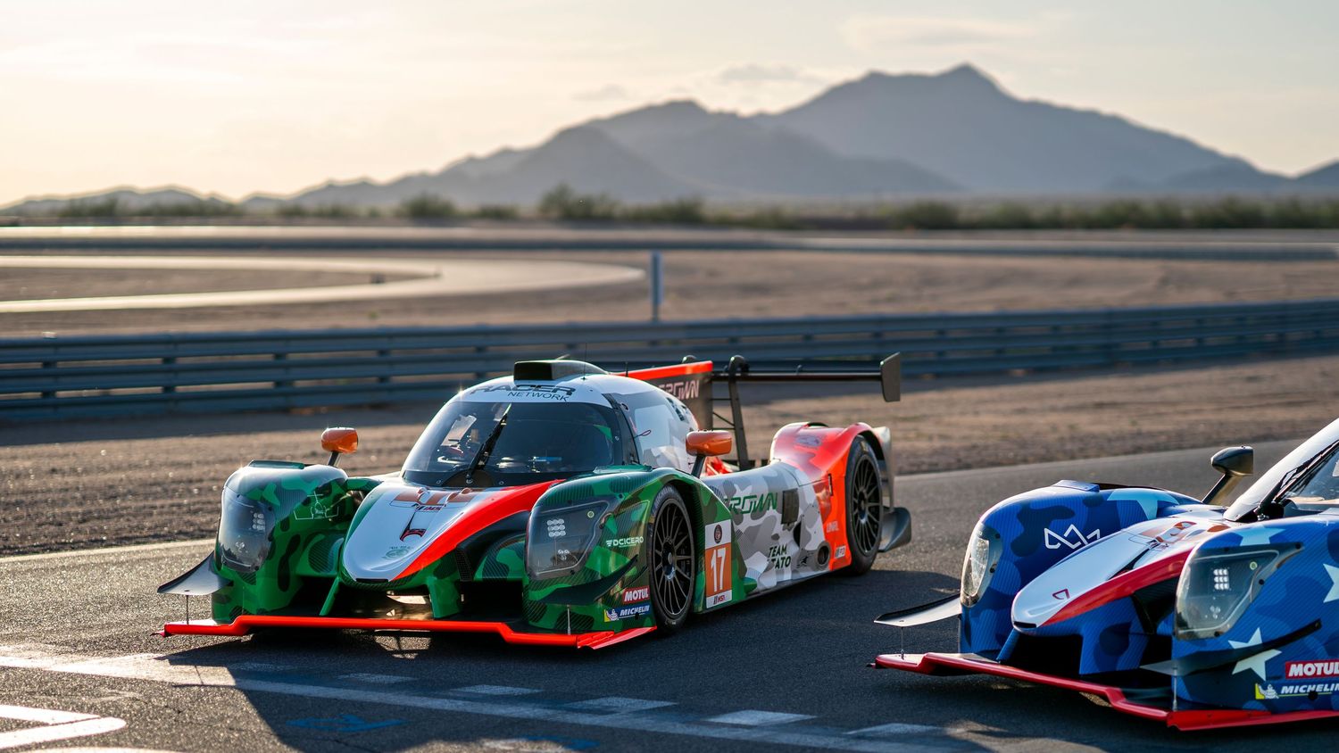 Motorsport racing cars lined up on a desert track with mountains in the background at sunset.