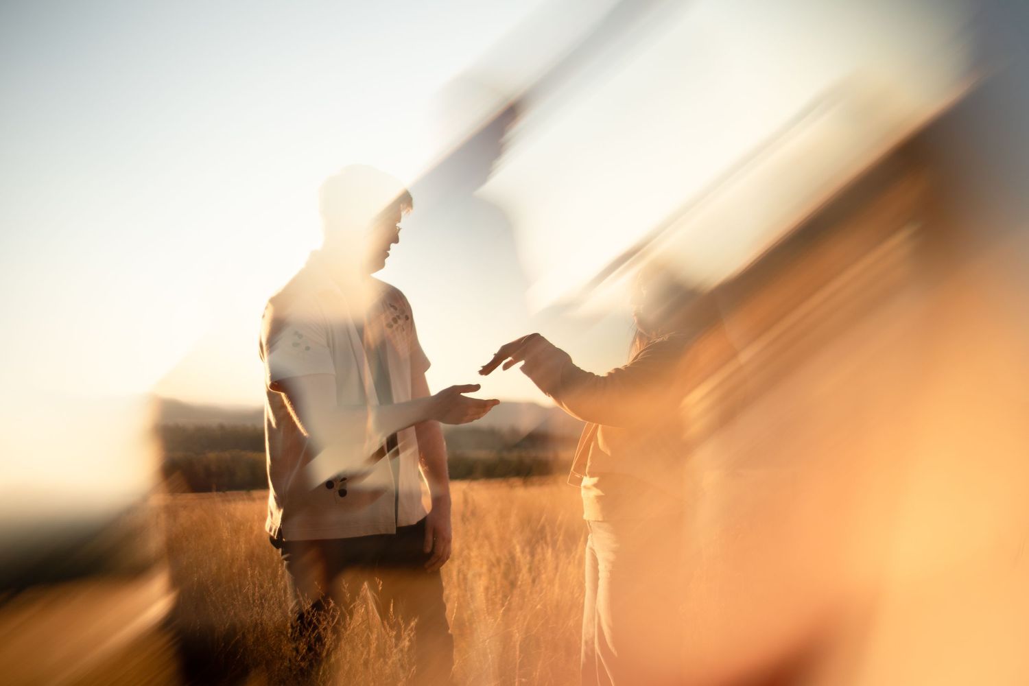 Two people stand facing each other in a golden field at sunset, reaching toward one another in a dreamy, blurred light-streaked photo.