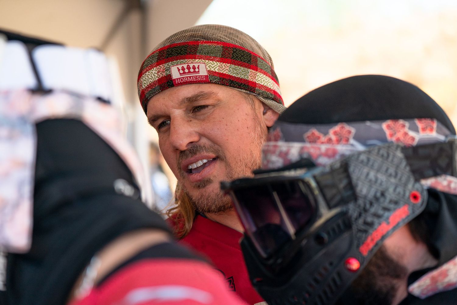 Man in colorful striped beanie holding paintball gear at outdoor event.