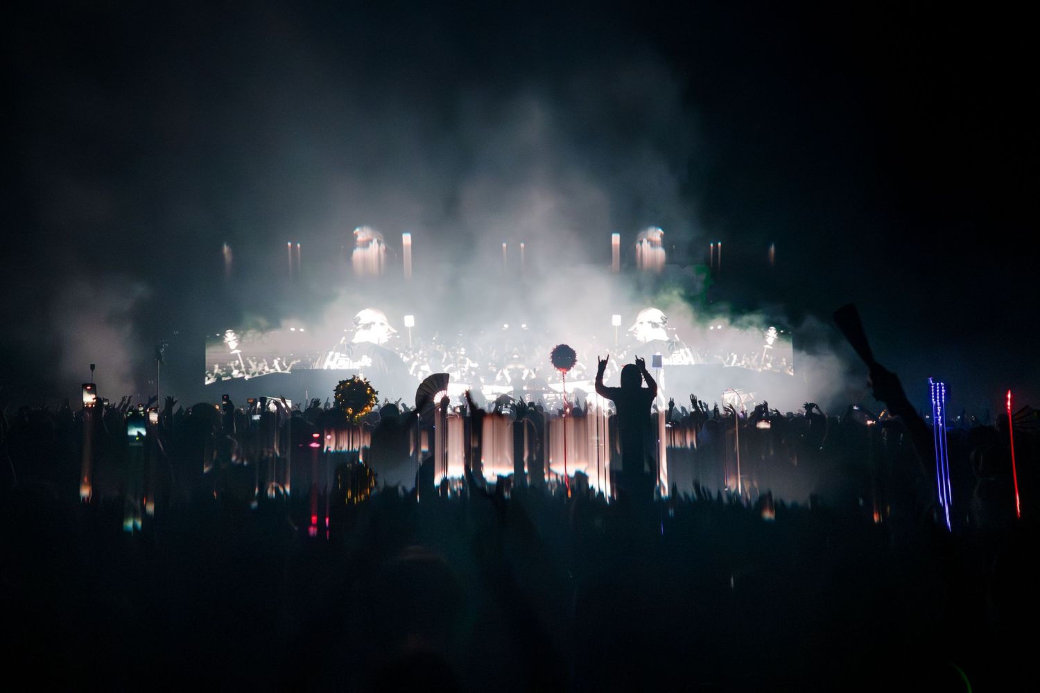 Crowd silhouettes at a massive outdoor music festival with dramatic fog and colorful stage lighting at night.