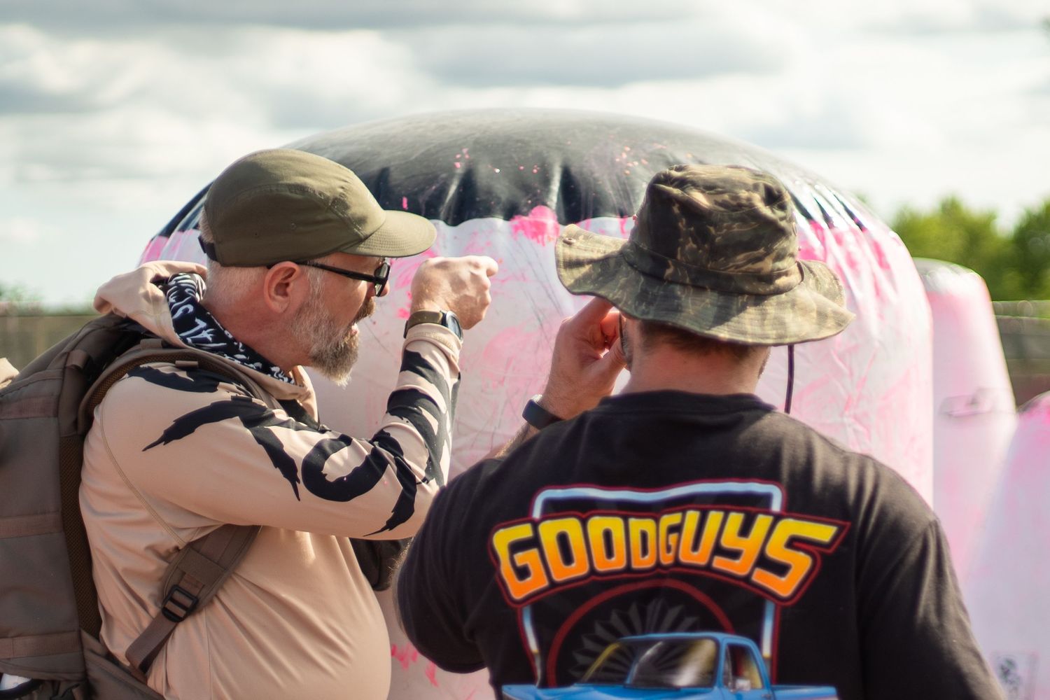 Two men in caps examine a pink inflatable structure outdoors at a Goodguys event.