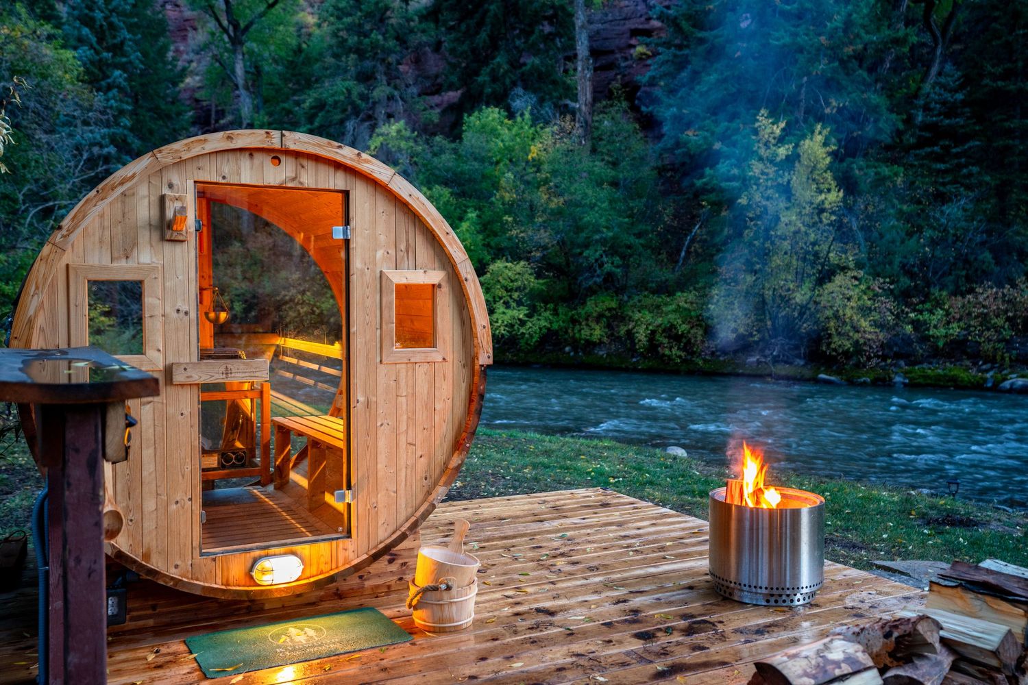 Cozy wooden barrel cabins with warm lighting nestled along a riverbank in a forested wilderness setting.