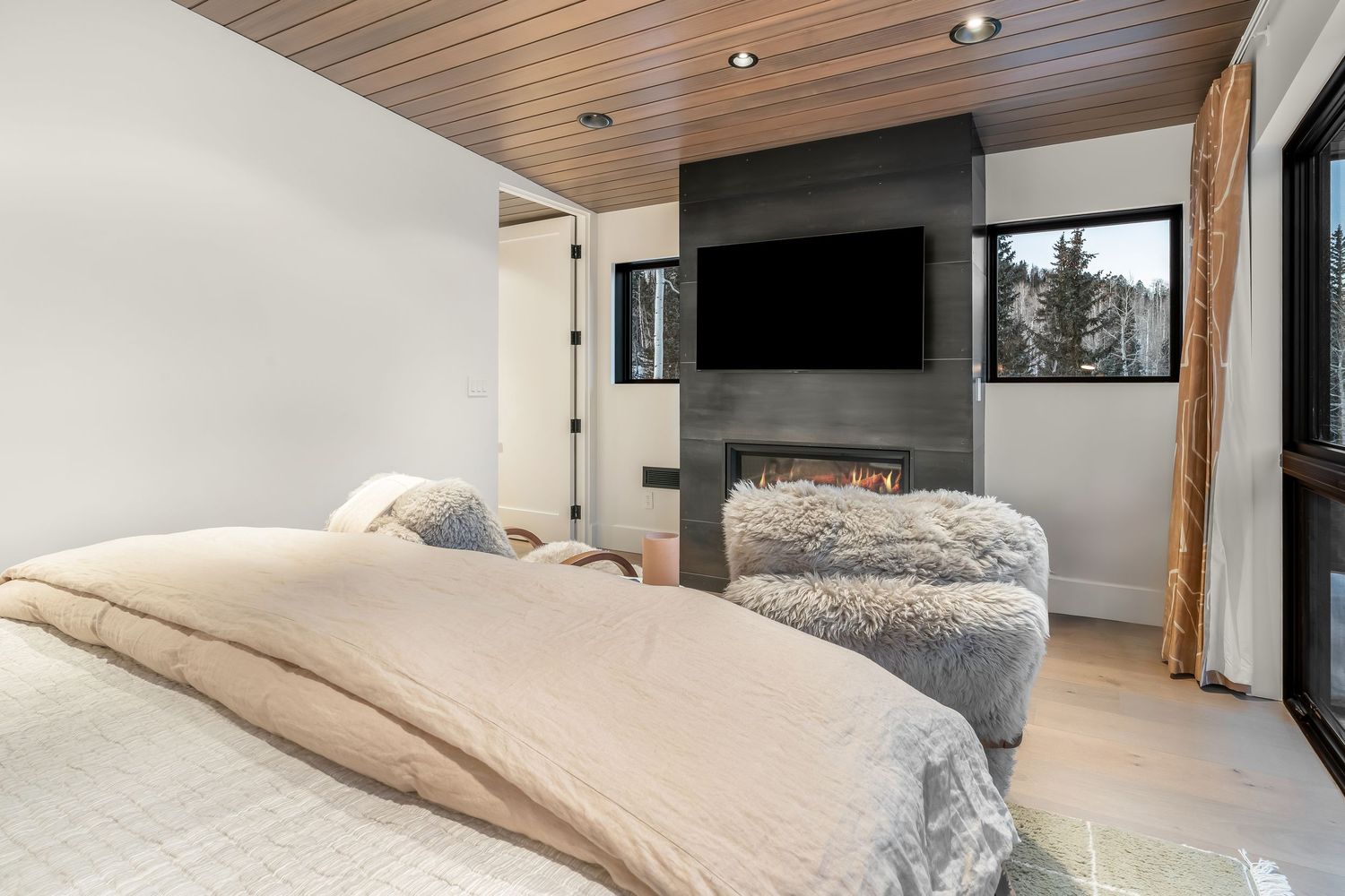 Modern bedroom with wood ceiling, wall-mounted TV, gray fireplace, and plush bedding in neutral tones.