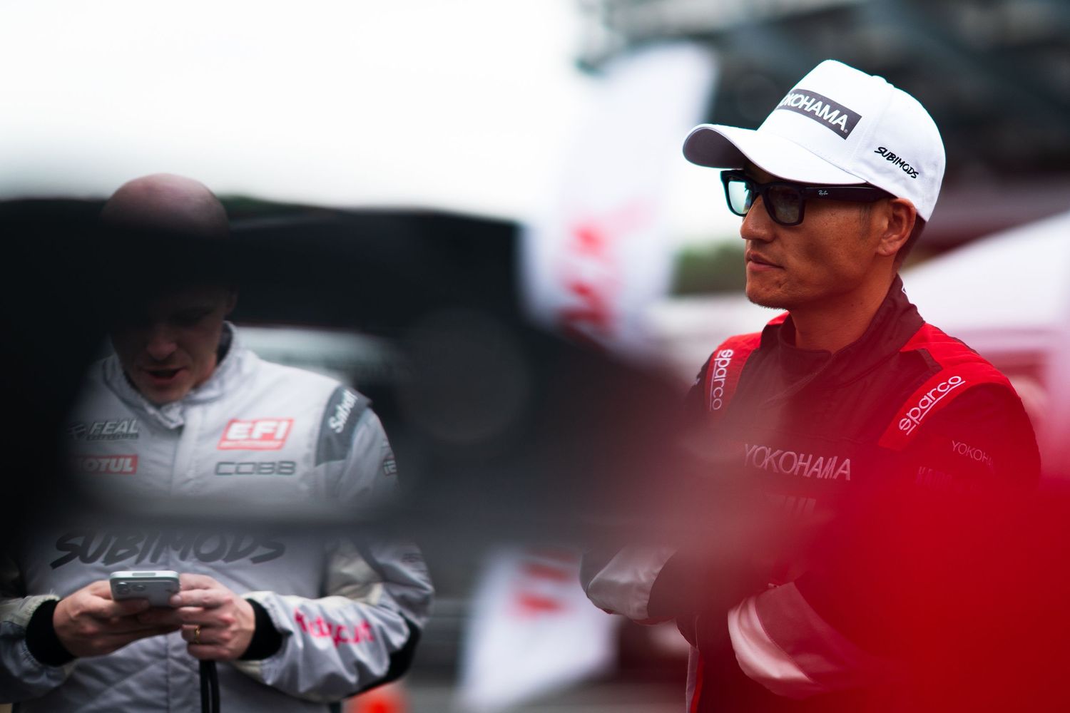 A motorsport driver wearing a Yokohama cap and sunglasses stands at a racing event.