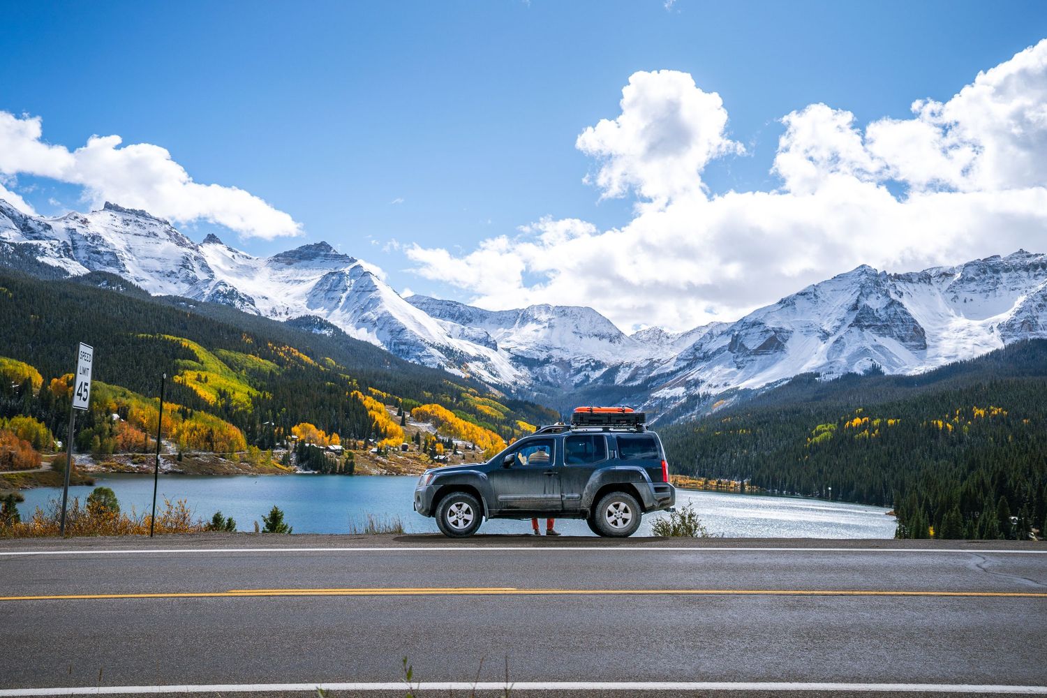 A panoramic mountain landscape with snow-capped peaks, autumn foliage, and a parked vehicle along a scenic road.