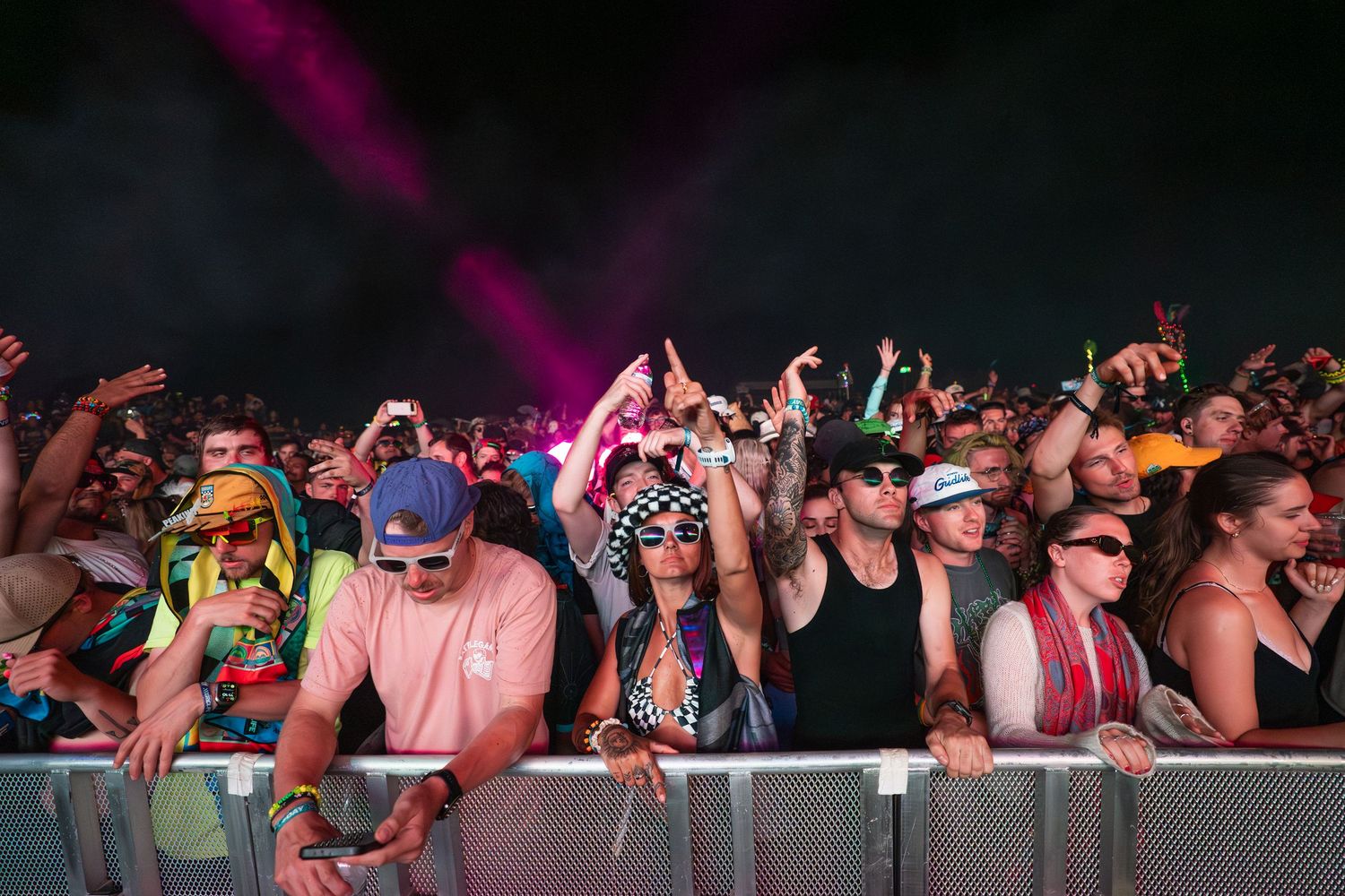 Excited crowd at an outdoor music festival at night with colorful stage lighting and fans cheering behind a barrier.