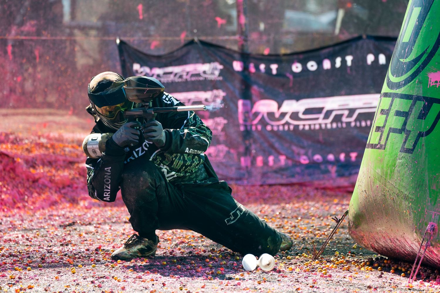 Paintball player crouching behind inflatable bunker during competitive match with colorful paint splatter.