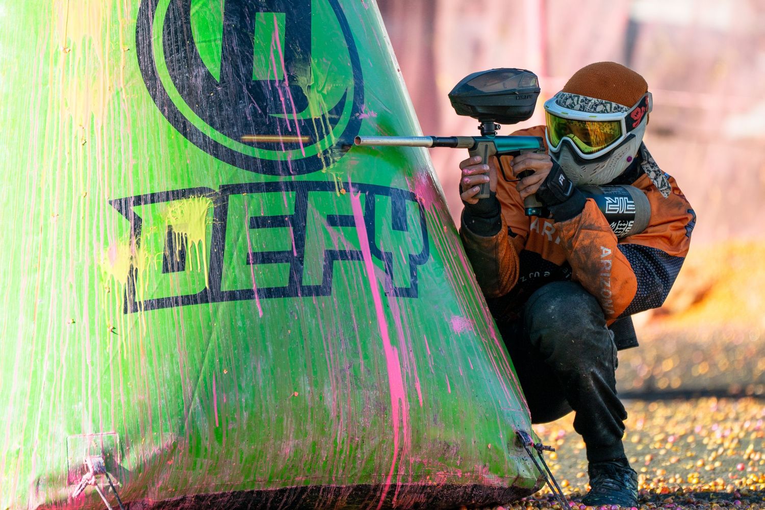 Paintball player in orange gear crouching behind a green DEFY bunker aiming their marker.