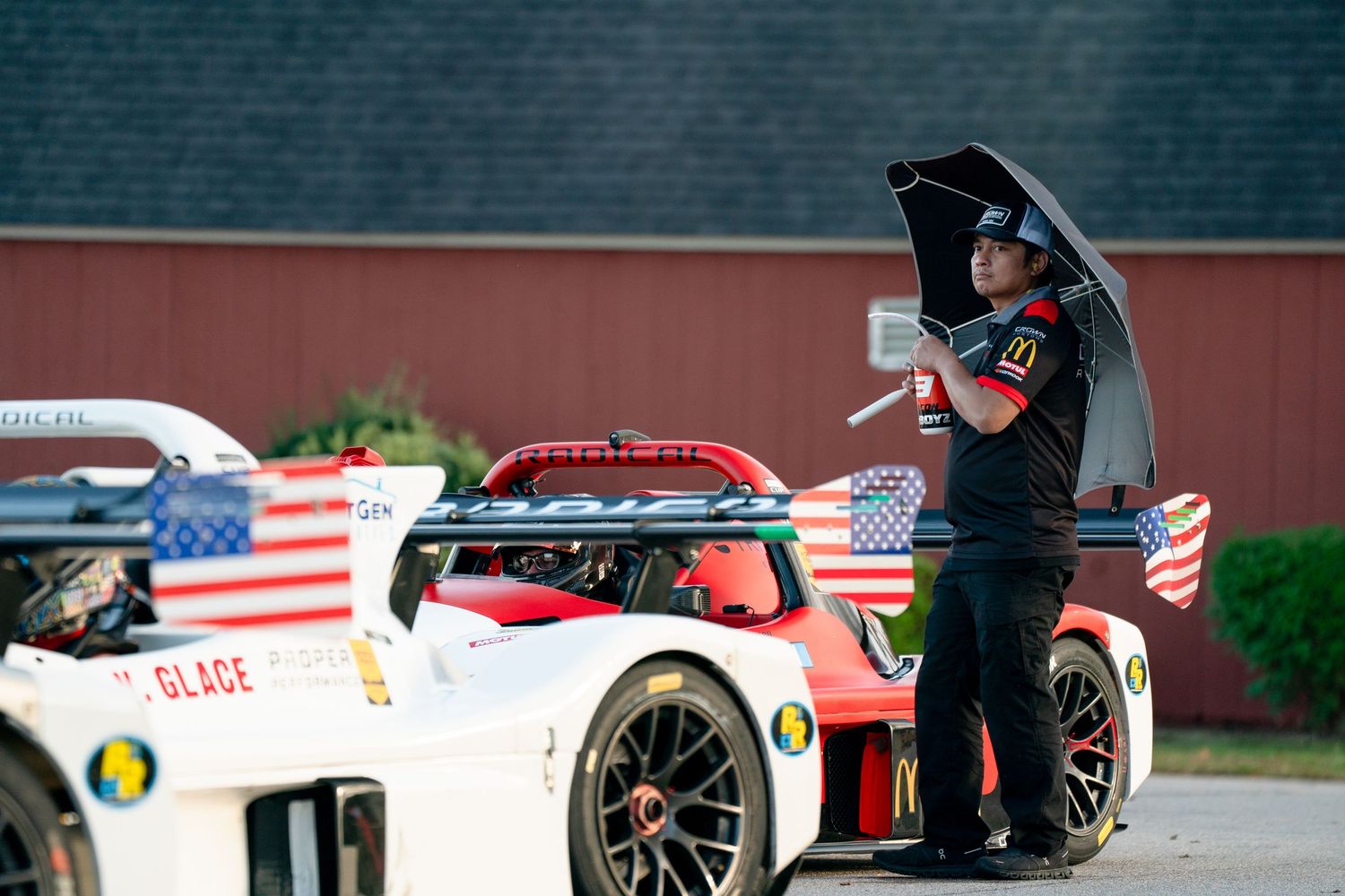 Race cars lined up in pit lane with American flags and team crews visible.