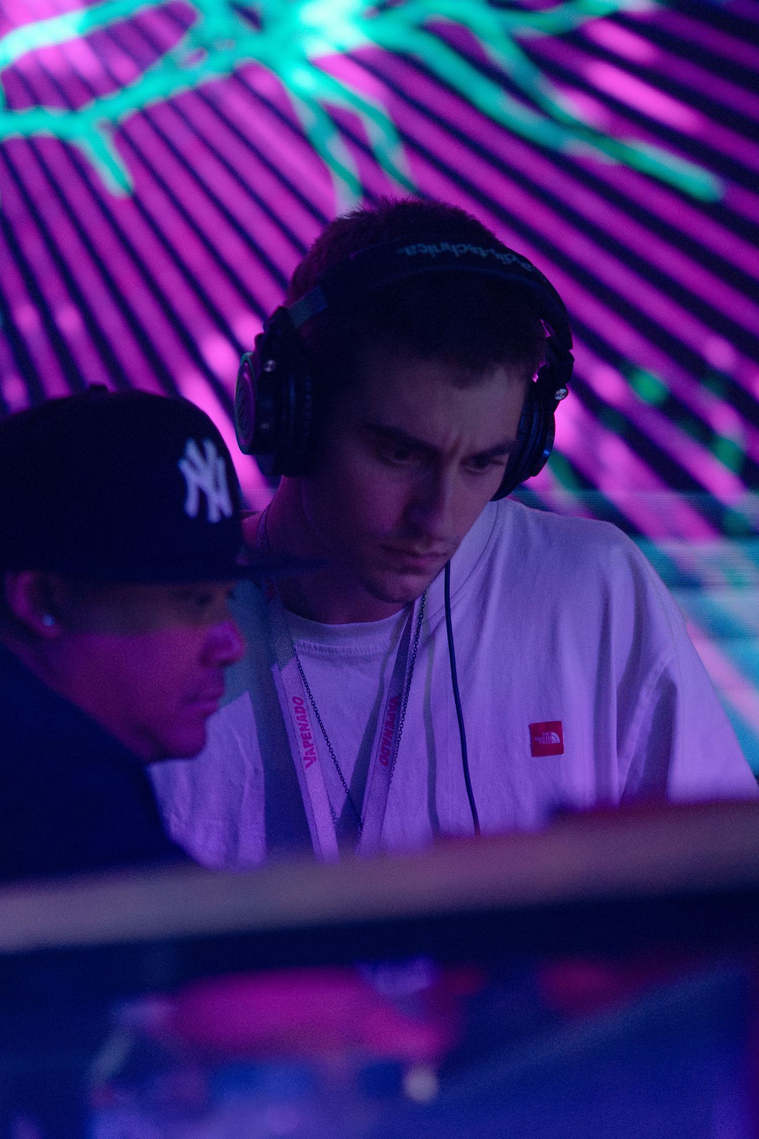 DJ performs in a dark club setting with neon purple and turquoise lighting while wearing headphones and a NY Yankees cap.