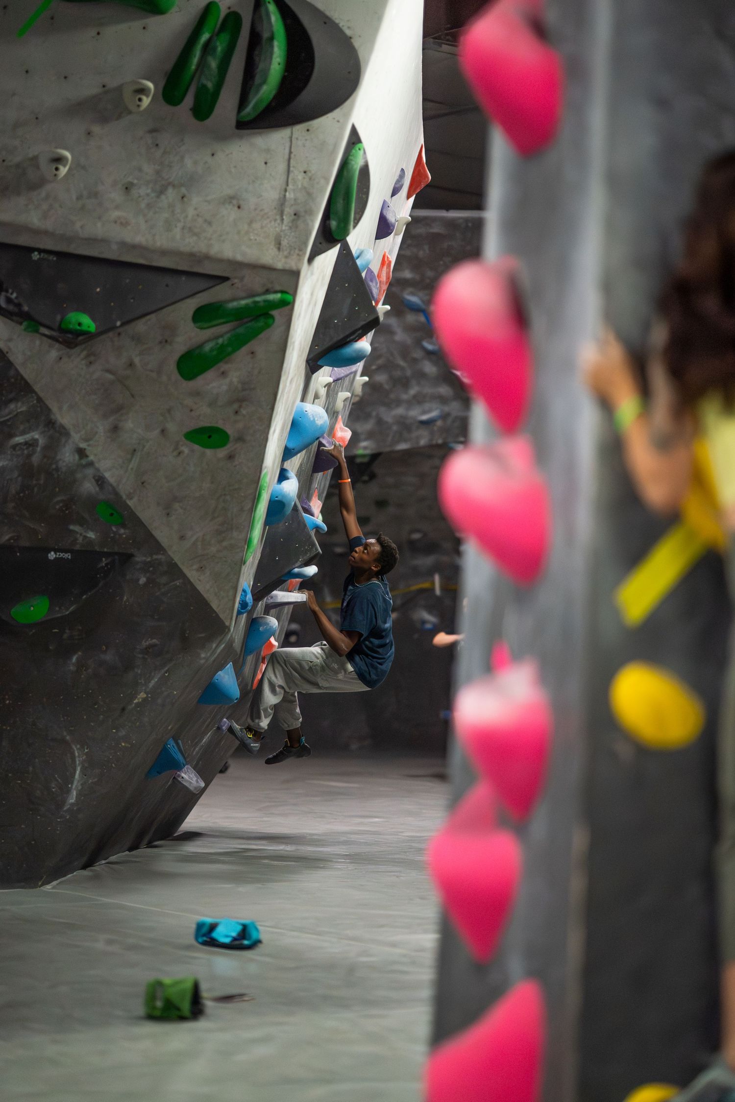 A climber in a blue shirt scales a gray indoor bouldering wall with colorful holds, framed by pink holds in the foreground.