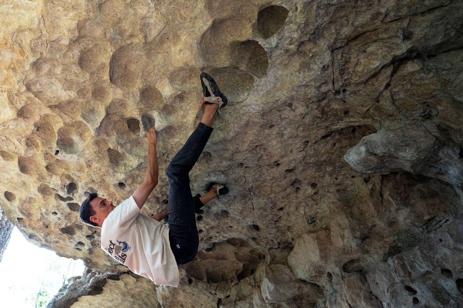 Rock climber in a white shirt and dark pants bouldering on an overhanging limestone wall with pocketed holes, one foot hooked high.