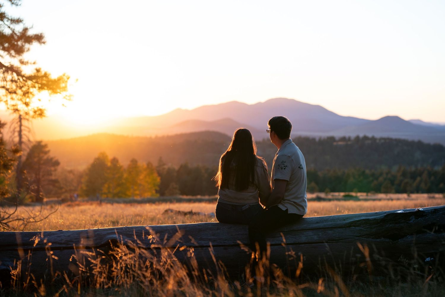 Couple sits close together on a wooden fence watching the sunset glow over distant mountains and a grassy valley.