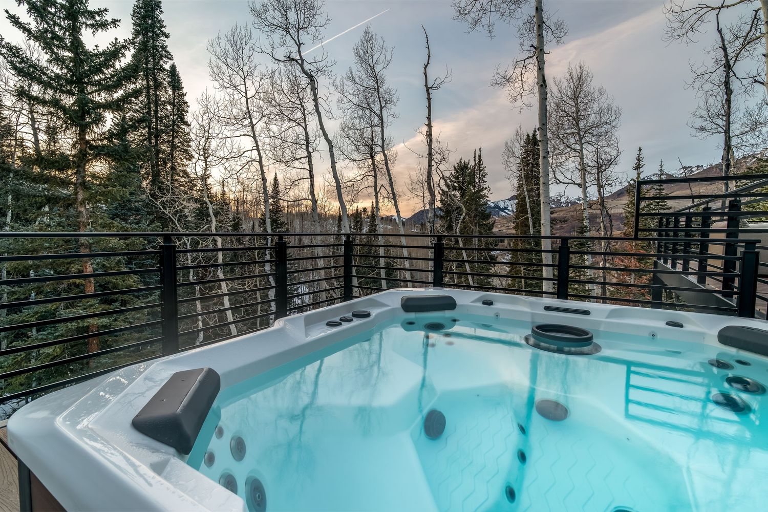 Illuminated hot tub on deck surrounded by aspen trees and mountain scenery.